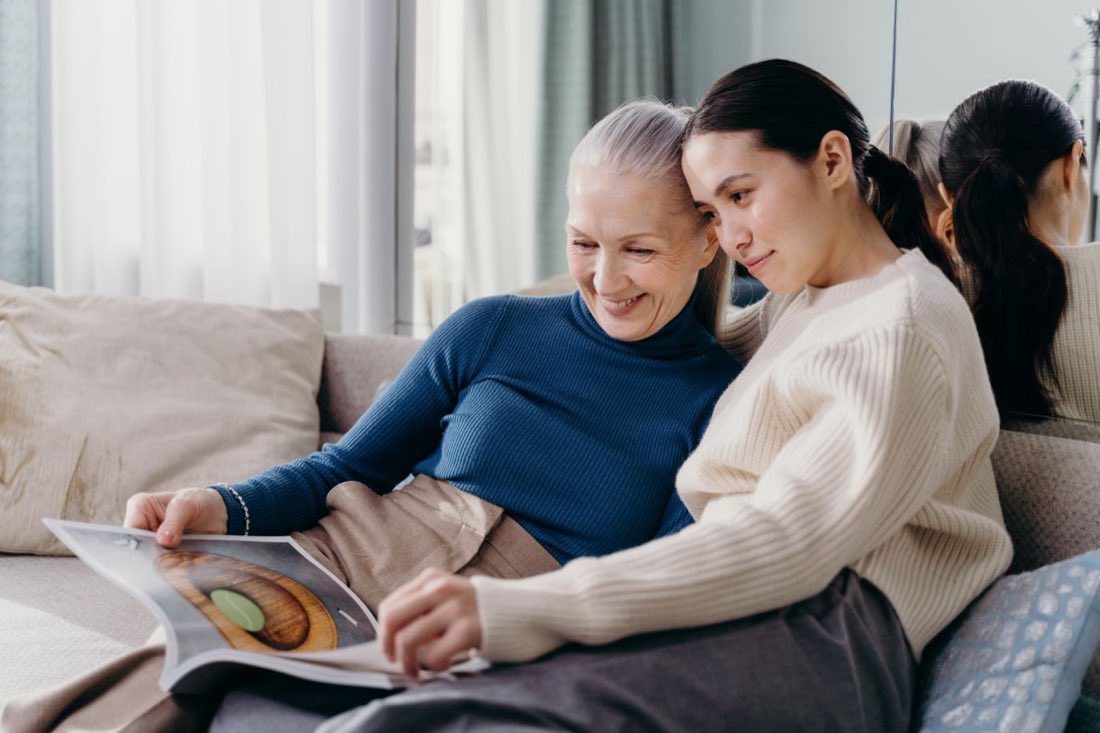 A senior mother and her adult daughter reading together on the couch at home